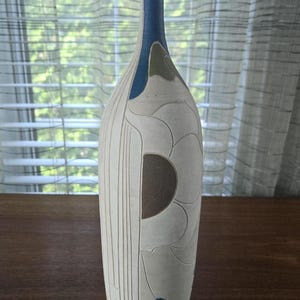 Puede incluir: Jarrón de cerámica blanco con un diseño abstracto en azul, marrón y verde. El jarrón tiene un cuello estrecho y una base ancha.