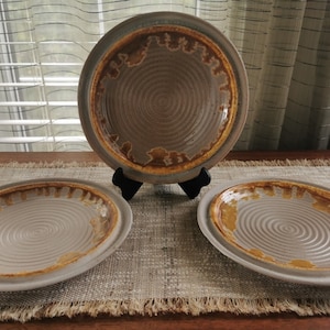 May include: Three round ceramic plates featuring a light gray center with concentric circular ridges and a wide, textured rim glazed in a mottled golden-brown finish. One plate is displayed upright on a black stand, while two lie flat.