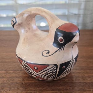 Op de afbeelding: Een klein, handbeschilderd keramiek vogeltje met een handvat. De vogel is wit met zwarte en rode geometrische patronen en een zwart en rood gezicht.