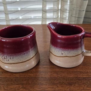 May include: A set of two ceramic creamer and sugar containers. The containers are a burgundy red color with a white and beige glaze.