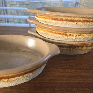 May include: Set of four beige ceramic baking dishes with brown and yellow glaze. The dishes are oval shaped and have a slightly raised rim.