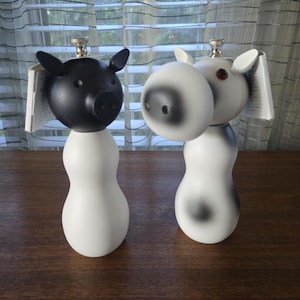 May include: Two decorative salt and pepper shakers. One is a black pig with a white body, the other is a white cow with black spots. Both have silver tops and are on a wooden surface.