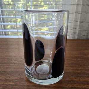 May include: A clear glass tumbler with a rounded base and a straight, cylindrical body. The glass is decorated with large, irregular black shapes. The tumbler sits on a dark brown wooden surface.