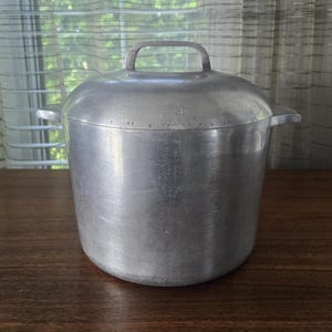 May include: A tall, cylindrical aluminium stockpot with a brushed metal finish. The pot features two small side handles and a matching domed lid with a central arched handle. The exterior surface shows a smooth, matte texture throughout the body.