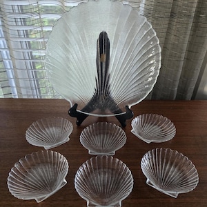 May include: A set of seven clear glass serving dishes shaped like seashells. The set includes a large serving platter and six smaller individual dishes.