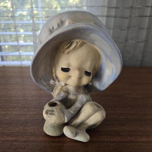 May include: A ceramic figurine of a child wearing a large, light blue hat. The child is seated, holding a small jug. The figurine has a serene expression with closed eyes and is painted in soft, muted tones.