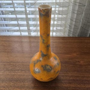 May include: A ceramic vase with a rounded base and a long, thin neck. The vase has a speckled orange and brown glaze with dark grey accents. The opening is a light beige colour.