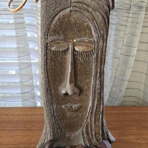 Könnte beinhalten: Handgefertigte, rechteckige Keramikskulptur eines ruhigen Gesichts mit geschlossenen Augen und langem Haar. Die Skulptur hat eine warme, gesprenkelte braune Farbe und ist auf einem Holzständer montiert. Oben ist ein Lederriemen befestigt.