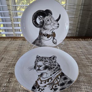 May include: Two white decorative plates with gold rims. One plate features a ram's head with large horns, a bow tie, and a pocket watch. The other plate has a leopard's head with a decorative collar. Both plates have black and white illustrations.