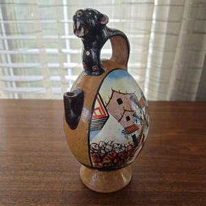 May include: A ceramic jug featuring a black panther head handle and a painted village scene. The jug is brown with a rounded body and a small spout. The village scene includes houses and trees.