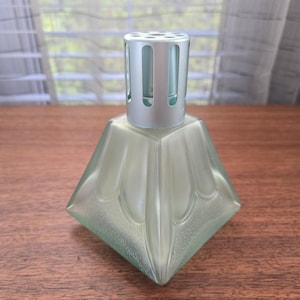 May include: A pale green, diamond-shaped fragrance lamp with a silver-coloured top. The glass lamp has a textured surface and a geometric design. The top has vertical slots for fragrance diffusion. The lamp sits on a wooden surface.
