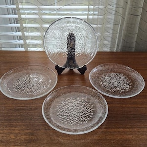 May include: A set of clear glass plates with a textured, bubble-like pattern. One plate is displayed on a black stand, while three others are arranged on a wooden surface. The plates are round and have a vintage aesthetic.