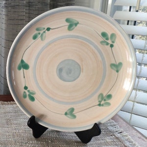 May include: A large, round ceramic plate with a light brown background and a blue centre circle. The plate is decorated with a green vine design around the edge.