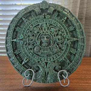 May include: A circular green wall plaque featuring a detailed relief of the Aztec sun stone calendar. The design includes a central face surrounded by concentric rings of intricate glyphs and symbols, supported by a silver metal display stand.