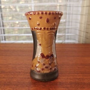 Puede incluir: Un jarrón de cerámica marrón y negro con un diseño de corona y rostro. El jarrón está decorado con puntos marrones y rojos.