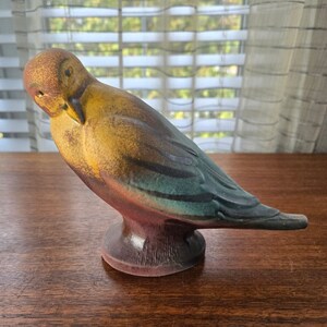 May include: A ceramic bird figurine with a yellow, green, and brown glaze. The bird is perched on a base and has its head tilted down.