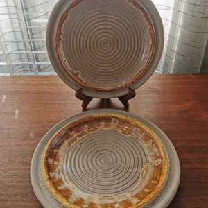 May include: Two ceramic plates with a white glaze and a brown rim. The top plate has a simple design, while the bottom plate has a more intricate design with a brown glaze on the rim.