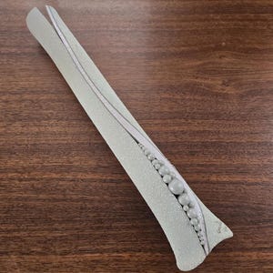 May include: A long, slender, light gray decorative object with a textured, granular surface. It features a curved, smooth white central ridge adorned with a row of graduated spherical beads that increase in size toward the wider base.