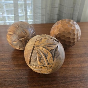 May include: Three wooden decorative spheres with unique carved designs. One sphere has a leaf pattern, another has a textured surface, and the third has a series of carved lines. The spheres are brown and sit on a wooden surface.