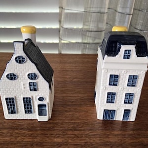 Op de afbeelding: Twee keramische huisvormige beeldjes met een witte baksteenstructuur en donkerblauwe accenten op de daken, ramen en deuren. Elk miniatuurgebouw is bekroond met een klein geel schoorsteenelement en toont gedetailleerde architecturale kenmerken.