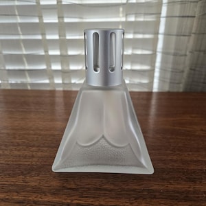 May include: A frosted glass aromatherapy diffuser with a silver metal top. The diffuser has a triangular base with a decorative scalloped edge. The top has vertical slots for fragrance diffusion. The diffuser sits on a wooden surface.