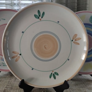 May include: A white ceramic dinner plate with a light blue, beige, and green floral design. The plate has a raised rim and a slightly textured surface.