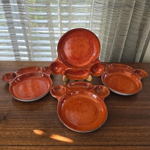 Può includere: Set di quattro piatti da portata in ceramica arancione. I piatti hanno un design unico con una grande area centrale e sezioni più piccole e arrotondate. La glassa ha un aspetto maculato. I piatti sono su una superficie di legno.