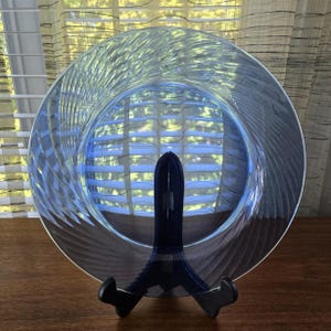 Può includere: Un piatto di vetro trasparente e circolare con un motivo a spirale, esposto su un supporto nero. I bordi del piatto presentano un motivo strutturato e la luce che lo attraversa crea una tonalità blu. Il piatto ha un diametro di circa 25 cm.