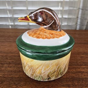 Könnte beinhalten: Eine Keramik-Schmuckdose mit einem Entenkopf-Deckel. Die Ente ist in realistischen Farben mit braunen, weißen und gelben Federn bemalt. Die Dose ist mit einem grünen Band und einer gelben und braunen Feldszene verziert.