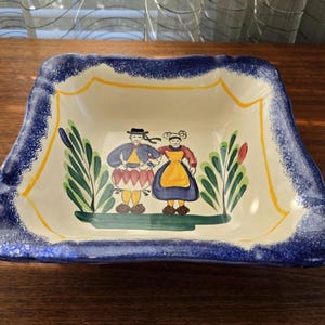 May include: Square ceramic serving dish with a hand-painted design. The dish features a blue rim, yellow accents, and a central image of two figures in traditional clothing. Green foliage surrounds the figures.