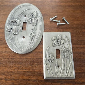 Puede incluir: Dos cubiertas decorativas de metal para interruptores de luz con diseños florales. Una es ovalada, la otra rectangular. Ambas tienen una abertura para un solo interruptor y están adornadas con flores y hojas de iris en relieve. También se ven varios tornillos.