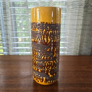 Puede incluir: Un jarrón de cerámica alto y cilíndrico con un esmalte moteado marrón y amarillo. El jarrón tiene una base redonda y lisa y una parte superior ligeramente acampanada.