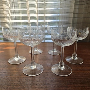 May include: Six clear crystal wine glasses with etched starburst patterns. The glasses have a rounded bowl, a slender stem, and a circular base. They are arranged on a dark wooden surface, with a window and sheer curtains in the background.
