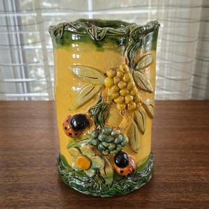 Puede incluir: Un jarrón cilíndrico de cerámica con base amarilla y borde verde. El jarrón está decorado con diseños en relieve de hojas, bayas, mariquitas y flores. Las mariquitas tienen manchas negras y las bayas son amarillas.