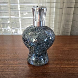 May include: A decorative glass bottle with a mosaic design in shades of gray and blue. The bottle has a silver-colored metal top. The bottle is sitting on a wooden surface.