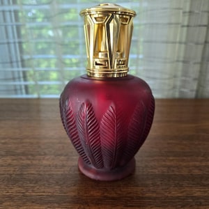 Puede incluir: Una lámpara de fragancia de vidrio esmerilado en color rojo intenso con un patrón de plumas en relieve en el cuerpo. El recipiente está rematado con una corona de metal dorado pulido con recortes geométricos y una tapa circular.