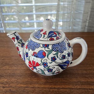 Puede incluir: Una tetera de cerámica blanca con tapa, asa y boquilla. La tetera está decorada con motivos florales azules, rojos y verdes. El diseño incluye lunares azules y flores estilizadas. La tetera está sobre una superficie de madera.