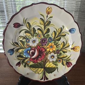 Op de afbeelding: Een wit keramiek bord met een gegolfde rand en een handgeschilderd bloemenontwerp. De bloemen zijn in tinten rood, geel, blauw, groen en paars.