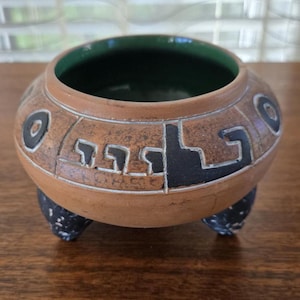 May include: A small, brown ceramic pot with black and white geometric designs. The pot has three black legs and a green glazed interior.