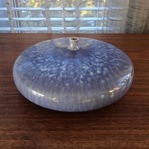 May include: A round, blue ceramic vase with a speckled pattern. The vase has a low profile with a small, cylindrical opening at the top. The colour is a mix of light and dark blues, creating a textured appearance. The vase is on a wooden surface.