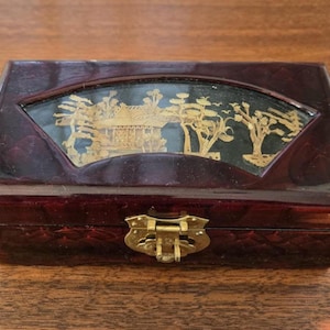 May include: A rectangular, dark burgundy jewellery box with a fan-shaped, clear window displaying a gold-coloured Asian landscape design. The box has a brass clasp and hinges, suggesting a vintage or antique style.