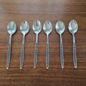 May include: Six silver spoons with oval bowls and long handles. The handles have a black decorative design. The spoons are arranged in a row on a wooden surface.