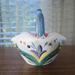 May include: A small, ceramic basket with a textured, off-white exterior. The basket features a blue handle and is decorated with colorful floral patterns in blue, green, yellow, and red. The basket sits on a wooden surface.