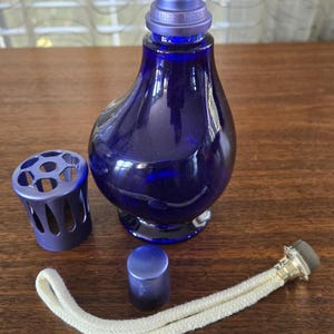 Pu&ograve; includere: Lampada a olio in vetro blu scuro smontata, con corpo bulboso. I componenti includono una gabbia di metallo, un piccolo tappo, uno stoppino e una parte superiore con la scritta "LAMPE BERGER". La lampada &egrave; su una superficie di legno.