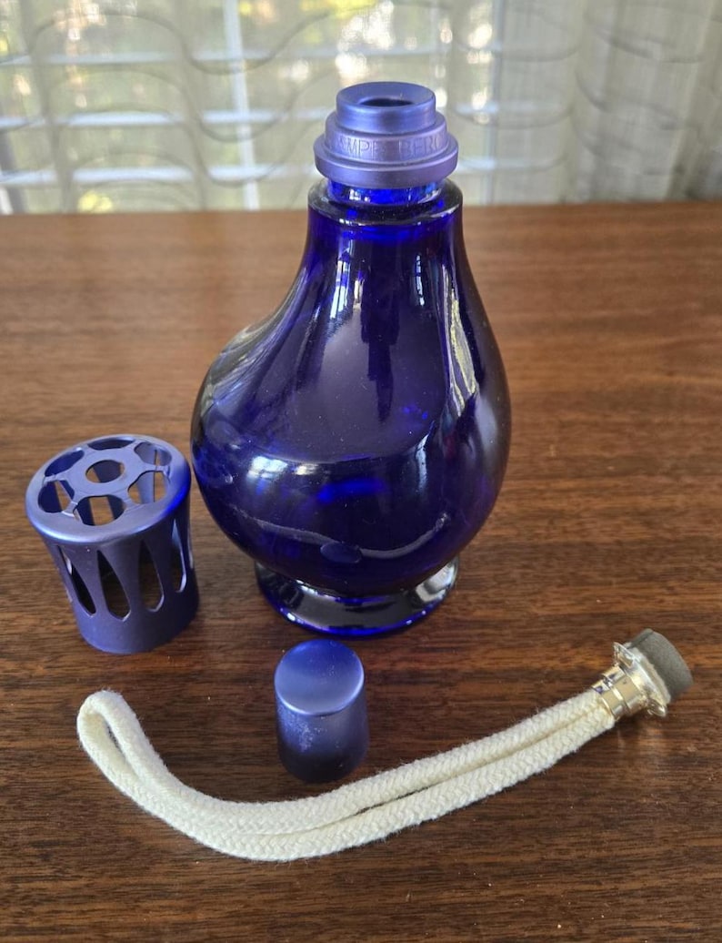 Pu&ograve; includere: Lampada a olio in vetro blu scuro smontata, con corpo bulboso. I componenti includono una gabbia di metallo, un piccolo tappo, uno stoppino e una parte superiore con la scritta "LAMPE BERGER". La lampada &egrave; su una superficie di legno.