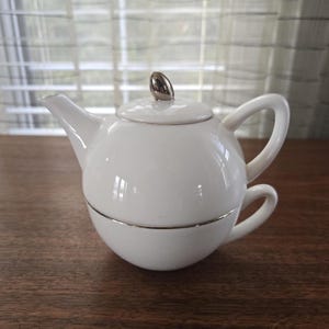 Puede incluir: Un juego de té individual de porcelana blanca. La tetera tiene un pomo plateado en la tapa y una banda plateada en el centro. La taza está integrada en el diseño, formando una unidad compacta. El juego está sobre una superficie de madera.
