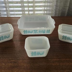 Puede incluir: Un juego de fuentes Pyrex blancas vintage con un diseño de gallo y floral turquesa. El juego incluye una fuente rectangular para hornear, una fuente cuadrada y dos fuentes pequeñas con tapa. Las fuentes están dispuestas sobre una superficie de madera oscura.