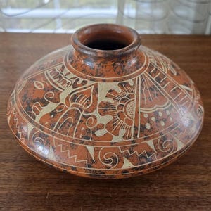 May include: A round, terracotta ceramic vase with a wide base and a narrow neck. The vase is decorated with a cream-coloured, etched design featuring floral and geometric patterns. The vase sits on a wooden surface.