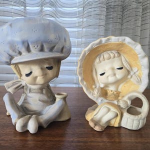 Könnte beinhalten: Zwei Keramikfiguren. Eine ist eine sitzende Figur mit blauer Haube und Latzhose. Die andere ist ein Mädchen mit gelber Haube, das einen Korb hält. Beide haben geschlossene Augen und sind in gedämpften Tönen bemalt.