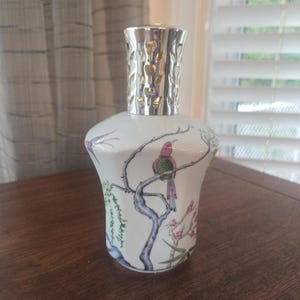 Puede incluir: Una lámpara de fragancia de cerámica blanca con acabado brillante, que presenta un diseño pintado a mano de un pájaro rosa y verde posado en una rama azul oscuro, rodeado de hojas de sauce verdes y flores rosas, con una tapa de metal plateado.